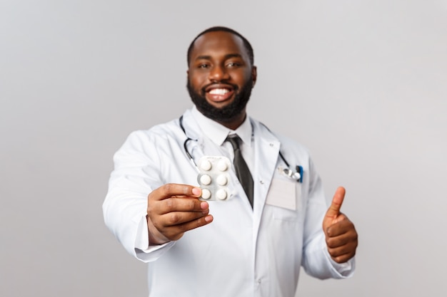 Portret zadowolonego, pewnego siebie afroamerykańskiego lekarza poleca pigułki, podaje pacjentom leki na receptę, trzyma pigułki, kciuk