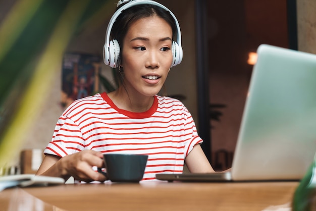 Portret Zadowolonej Azjatyckiej Kobiety Słuchającej Muzyki Przez Słuchawki Podczas Korzystania Z Laptopa W Kawiarni W Pomieszczeniu