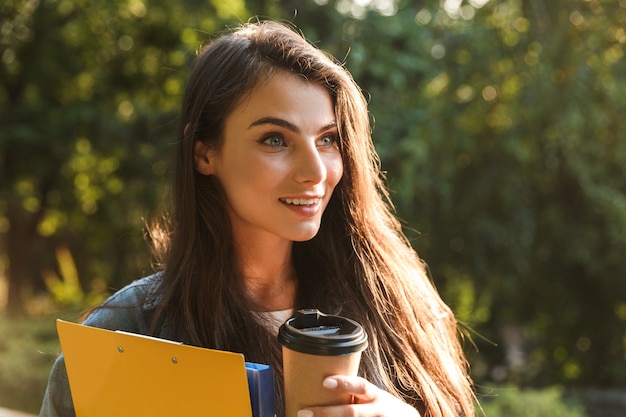 Portret zadowolonej brunetki uśmiecha się i pije kawę na wynos podczas spaceru z książkami w zielonym parku w letni dzień