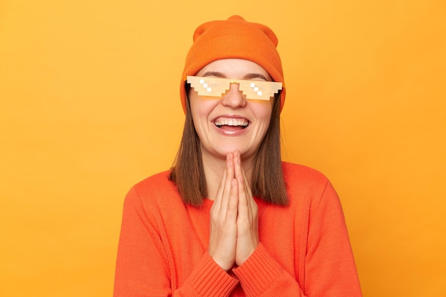 Portret zadowolonej hipsterki w pomarańczowym swetrze, kapeluszu i okularach w stylu minecraft, pozującej się na białym tle na żółtym tle, trzyma dłonie w modlitewnym geście błagalnym