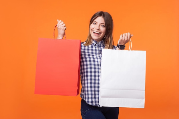 Portret zadowolony kupujący, brunetka kobieta z uroczym uśmiechem w dorywczo kraciastej koszuli stojący pokazujący torby na zakupy, chwalący się zakupem do aparatu. strzał studio w pomieszczeniu na białym tle na pomarańczowym tle