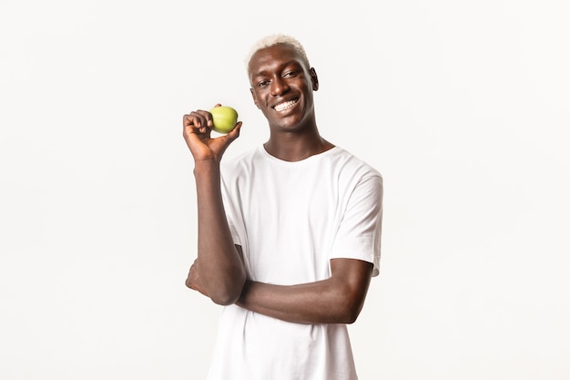 Portret zadowolony, przystojny i wesoły Afroamerykanin blondyn, pokazując jabłko i uśmiechając się z doskonałymi białymi zębami, stojąc w studio.
