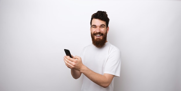 Portret zadziwiający brodaty mężczyzna w białym koszulki mienia smartphone i patrzeć kamerę