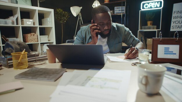 Portret zajętego menedżera afro piszącego notatki na dokumentach w pobliżu laptopa w biurze
