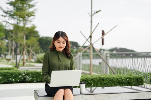 Portret zajęty sprzedaży kobieta siedzi w parku biznesu i za pomocą swojego laptopa podczas pisania tekstu na telefonie komórkowym. Kobieta pracuje online.