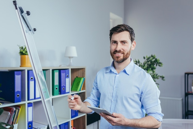 Portret założyciela firmy, który odniósł sukces, mężczyzna patrzy w kamerę i uśmiecha się do białej tablicy, aby zapisać notatki, trzymając tablet, opisuje strategię biznesową