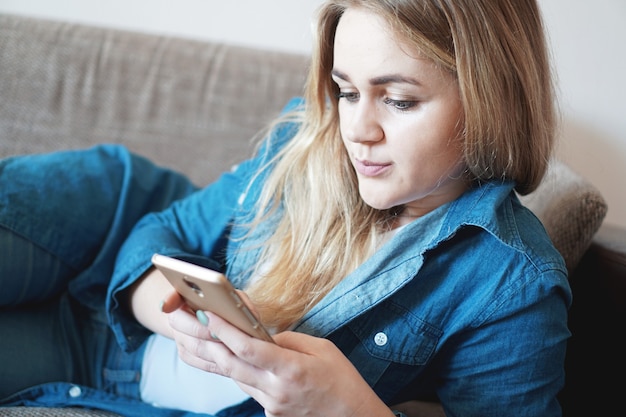 Portret zamyślonej kobiety leżącej na kanapie poduszki, trzymając w rękach inteligentny telefon, patrząc na ekran