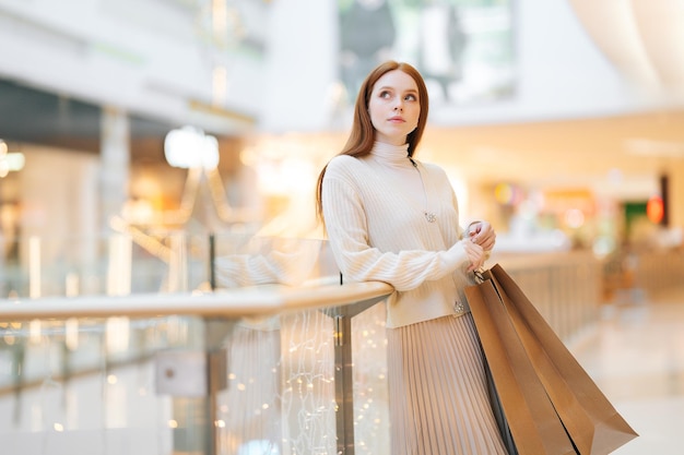 Portret zamyślony całkiem młoda kobieta trzyma w rękach zakupy papierowe torby z zakupem stojący w centrum handlowym z świątecznym wnętrzem, odwracając wzrok, rozmazane tło.