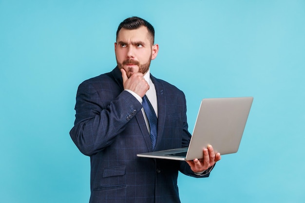 Portret zamyślony inteligentny biznesmen ubrany w ciemny garnitur służbowy, trzymający laptopa i myślący o nowym projekcie, trzymając podbródek Kryty studio strzał na białym tle na niebieskim tle