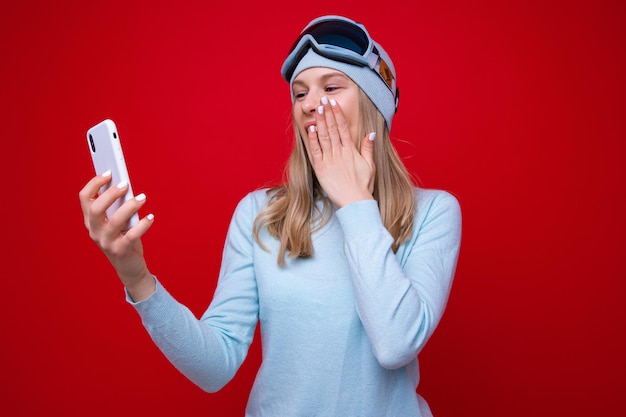 Portret zaskoczonej młodej kobiety w swetrze i goglach narciarskich z telefonem