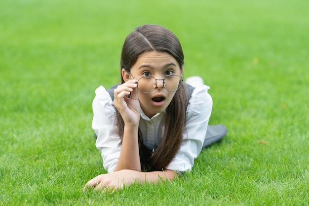 Portret zaskoczonej nastoletniej dziewczyny dostosowującej okulary leżącej na trawie po szkole niespodzianka Nastoletnia dziewczyna