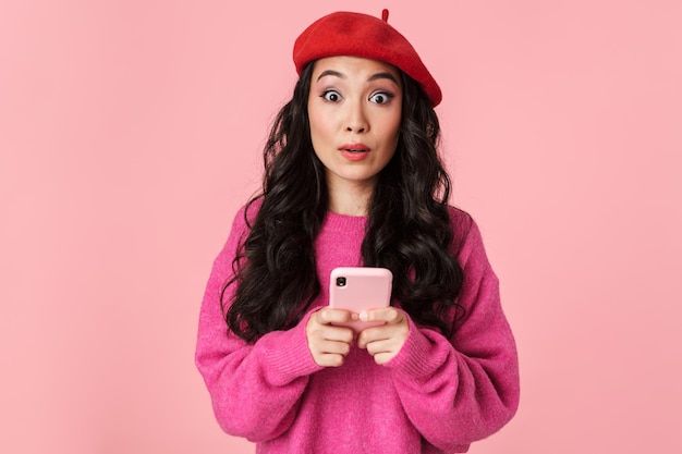 Portret zaskoczonej pięknej azjatyckiej dziewczyny w berecie, wyrażającej podziw i trzymającej smartfona odizolowanego na różowo