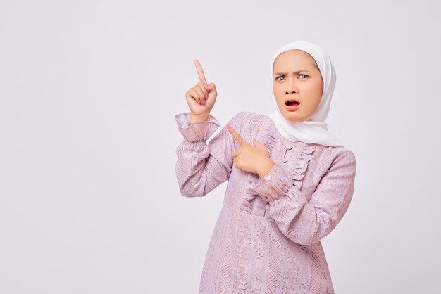 Portret zaskoczonej pięknej młodej azjatyckiej muzułmanki noszącej hidżab i fioletową sukienkę wskazującą palcem na wolne miejsce na reklamę odizolowaną na tle białego studia