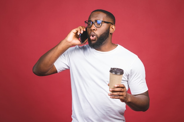 Portret Zaskoczony Przystojny Mężczyzna Afroamerykanów Z Telefonu Komórkowego I Zabrać Filiżankę Kawy. Pojedynczo Na Czerwonym Tle.