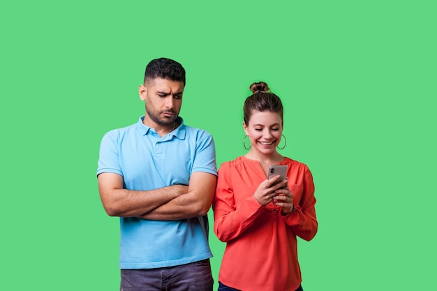 Portret zazdrosnego chłopaka stojącego ze skrzyżowanymi ramionami, patrzącego podejrzliwie na kobietę, która zdradza za pomocą telefonu i uśmiecha się szczęśliwie niewierne relacje na białym tle na zielonym tle strzał w studio