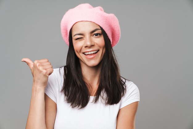 Portret zbliżenie piękna ładna kobieta w podstawowym t-shirt i beret mrugając, wskazując palcem na bok na lato na białym tle nad szarą ścianą