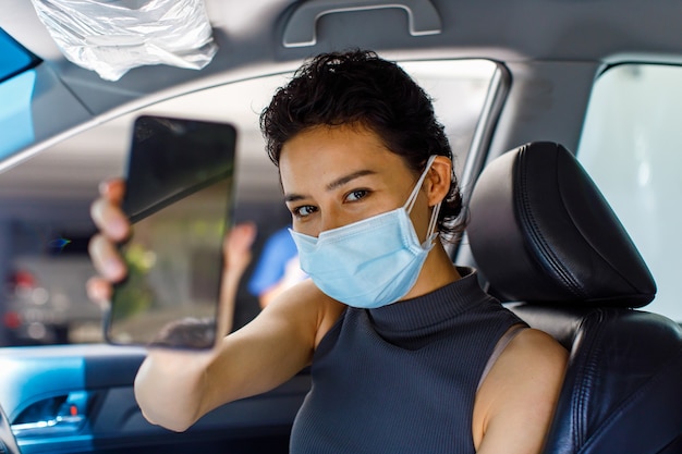 Portret zbliżenie strzał kobiety noszącej maskę siedzącą w samochodzie w przejeździe przez linię do szczepień przeciwko koronawirusowi, trzymając pusty czarny telefon komórkowy na kopię miejsca na niewyraźnym pierwszym planie.