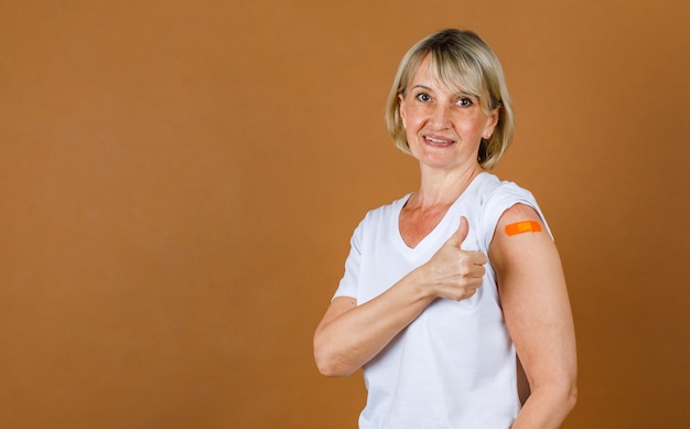 Portret Zbliżenie studio strzał kaukaski starszy blond kobieta pacjenta spojrzeć na kamery Pokaż kciuk na pomarańczowy bandaż tynk na jej ramieniu po otrzymaniu szczepienia przeciwko koronawirusowi.