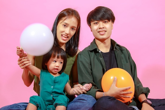 Portret zbliżenie studio strzał młodej szczęśliwej azjatyckiej rodziny ojca matki i małej słodkiej córeczki dziewczynka siedzi na podłodze razem uśmiechnięte spojrzenie na kamerę trzymającą kolorowe balony na różowym tle.