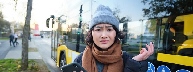 Portret zdezorientowanej Azjatki stojącej na przystanku autobusowym, trzymającej telefon komórkowy i wyglądającej na zszokowaną i zdenerwowaną