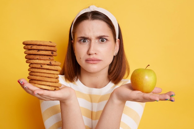 Portret zdezorientowanej kobiety w pasiastej koszuli i opasce do włosów patrzącej w kamerę ze zdziwieniem, trzymającej ciasteczka i jabłko, nie wie, co wybrać, pozując na białym tle na żółtym tle