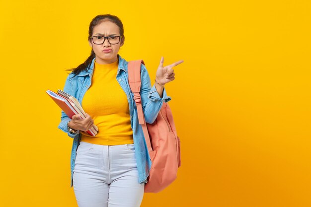 Portret Zdezorientowanej Młodej Studentki Azjatyckiej Kobiety W Dżinsowych Ubraniach Z Plecakiem Trzymającym Notatnik I Wskazującym Palcem Na Miejsce Na Kopię Na Białym Tle Na żółtym Tle