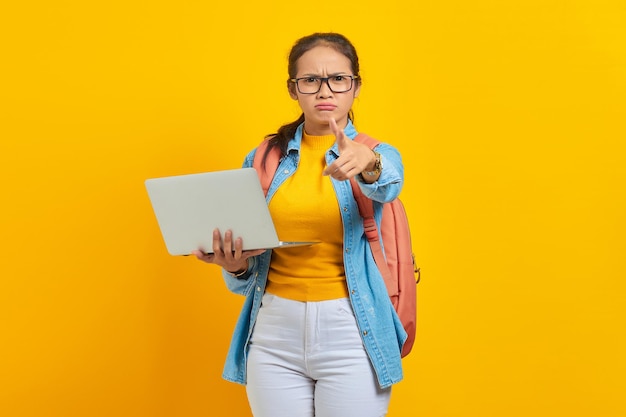 Portret zdezorientowanej młodej studentki azjatyckiej kobiety w zwykłych ubraniach z plecakiem za pomocą laptopa i wskazując na aparat palcem na białym tle na żółtym tle Edukacja w koncepcji uniwersytetu
