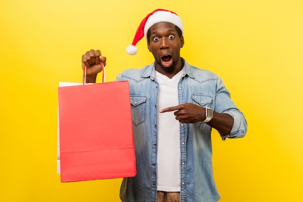 Portret zdziwionego przystojnego młodzieńca w santa hat i dżinsowej koszuli stojącej na co dzień, wskazującego na torbę na zakupy, zszokowanego bożonarodzeniowymi zniżkami. strzał w studio na białym tle na żółtym tle