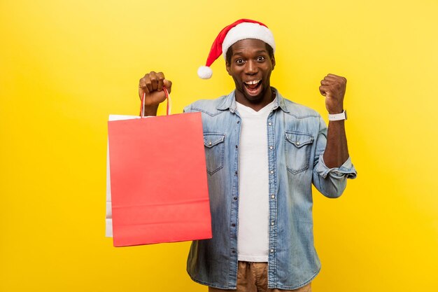 Portret zdziwionego szczęśliwego mężczyzny w santa hat i casualowej dżinsowej koszuli stojącej z podniesioną ręką pokazującą tak gest trzymający torby podekscytowany zakupami w studio strzał na białym tle na żółtym tle