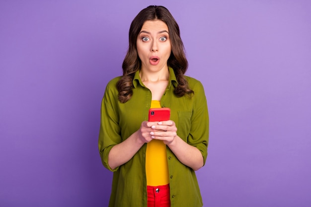 Portret zdziwionej dziewczyny używa telefonu komórkowego pod wrażeniem informacji z mediów społecznościowych, gapi się na osłupienie, nosi dobrze wyglądające ubrania odizolowane na fioletowym tle