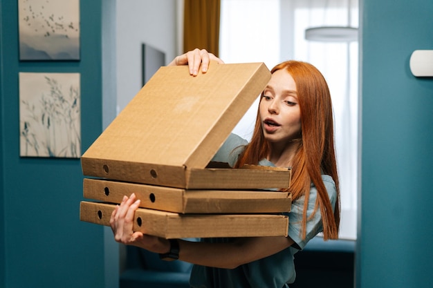 Portret zdziwionej podekscytowanej młodej kobiety, która otwiera pudełko gorącej pizzy stojącej w drzwiach do mieszkania, zaglądając do środka