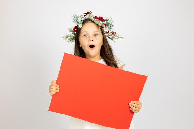 Portret zdziwionej, zszokowanej dziewczyny z otwartymi ustami w świątecznym wieńcu trzymającym czerwony pusty transparent z...