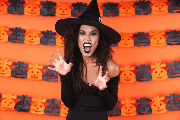 Portret zły czarownica dziewczyna w czarnym kostiumie halloween krzyczy i drapie się na pomarańczowej ścianie dyni