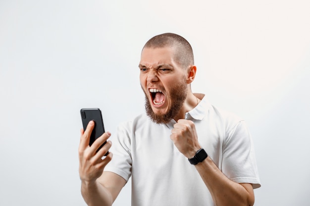 Zdjęcie portret zły przystojny brodaty mężczyzna w białej koszulce trzyma w ręku smartfon