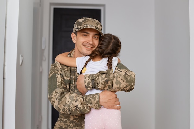 Portret żołnierza w mundurze kamuflażowym i czapce pozuje z córką, pozuje do tyłu i przytula swojego uroczego tatę, nie chce, żeby poszedł do wojska
