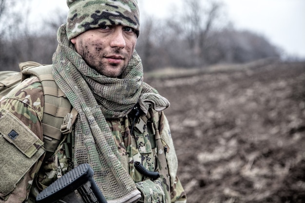 Portret żołnierza, współczesnego kombatanta z brudną twarzą, replika broni palnej, w mundurze maskującym, czapce i pelerynie maskującej na szyi, stojącego na polu, niosącego plecak w marszu