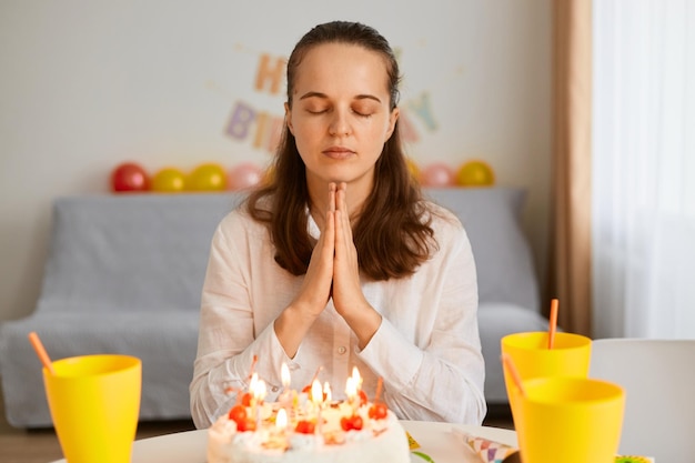 Portret zrelaksowanej kobiety o ciemnych włosach, siedzącej przy stole i trzymającej ręce razem, modlącej się z zamkniętymi oczami, świętującej wakacje samotnie pozujące w urządzonym świątecznym pokoju