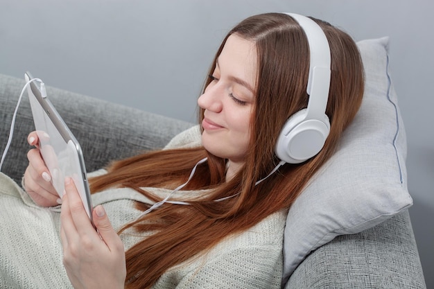 Portret zrelaksowanej kobiety ze słuchawkami, siedzącej na kanapie i słuchającej muzyki na cyfrowym tablecie