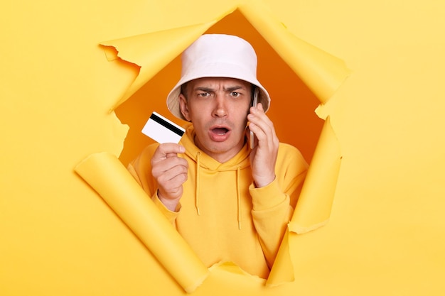 Portret zszokowanego przestraszonego mężczyzny noszącego bluzę z kapturem i panamę, przedzierając się przez rozdartą w żółtej ścianie papieru, stojącego rozmawiającego na telefonie komórkowym i pokazującego plastikową kartę kredytową