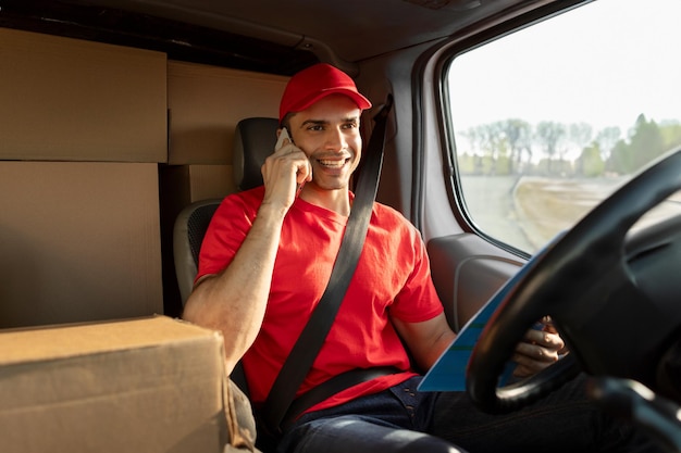 positive young courier man calling clients while driving van full of delivery boxes with parcel