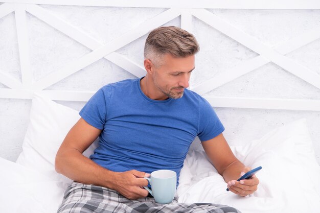 Posiwiały mężczyzna wysyłający SMS-y na telefon komórkowy podczas picia kawy w łóżku Wysyłanie wiadomości za pomocą aplikacji mobilnej