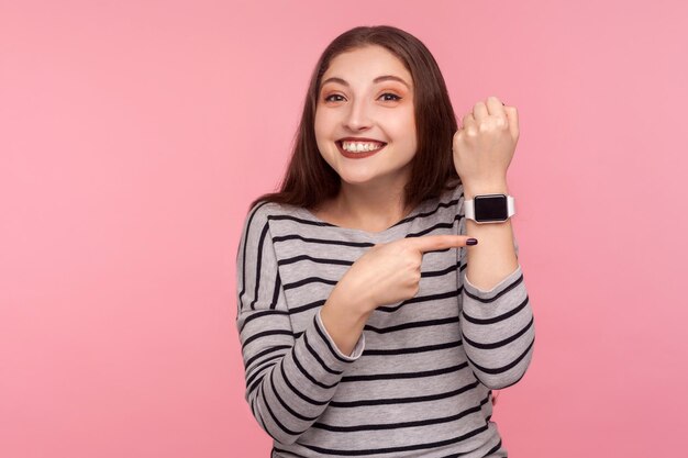 Pośpiesz się. Portret pozytywnej kobiety w pasiastej bluzie, wskazującej smartwatch na jej nadgarstku i patrzącej w kamerę z uśmiechem zębów, martwiące się o późny czas. studio strzał na białym tle na różowym tle