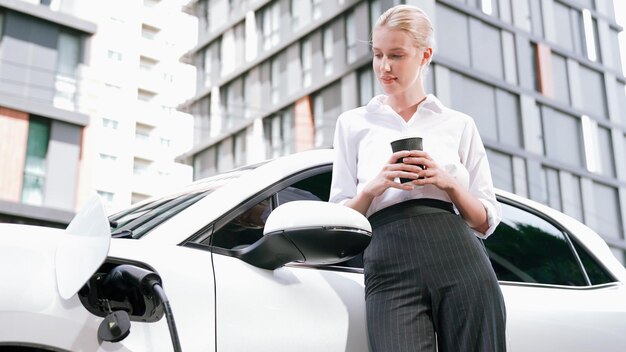 Postępowa bizneswoman opierająca się na samochodzie elektrycznym i stacji ładującej przed jazdą po centrum miasta Przyjazny dla środowiska samochód EV zasilany zrównoważoną i czystą energią