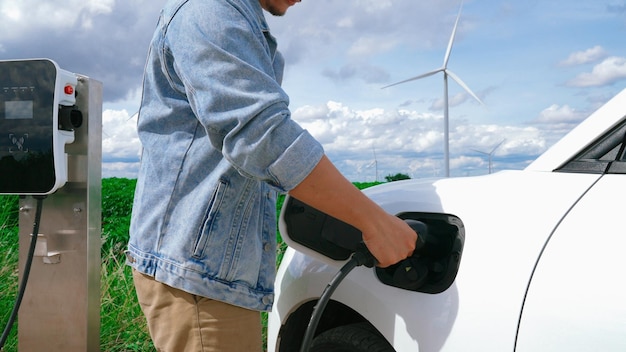 Postępowy człowiek ze swoim samochodem elektrycznym Samochód EV ładujący energię ze stacji ładowania na zielonym polu z turbiną wiatrową jako koncepcja przyszłej zrównoważonej energii Pojazd elektryczny z generatorem energii
