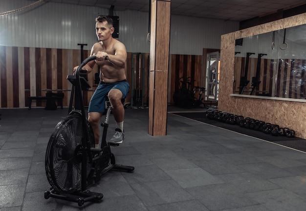 Potężny, umięśniony sportowiec z nagim torsem trenuje na rowerze powietrznym w nowoczesnej siłowni. Trening funkcjonalny. Ćwiczenia cardio. Koncepcja zdrowego stylu życia