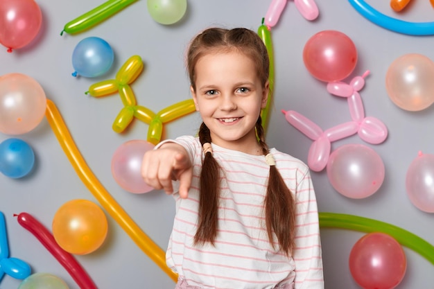 Potrzebuję cię Uśmiechnięta śliczna dziewczynka z warkoczami wskazującymi na kamerę, zapraszająca na swoje przyjęcie urodzinowe stojąca przy szarej ścianie ozdobionej kolorowymi balonami