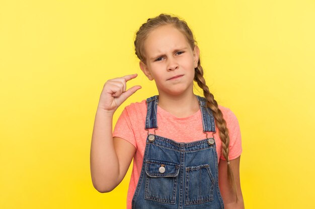 Potrzebujesz więcej. Portret zdenerwowanej dziewczynki w dżinsowych kombinezonach robi mały gest i patrzy z niezadowoloną błagalną miną, pokazując minimum. studio strzał na białym tle na żółtym tle