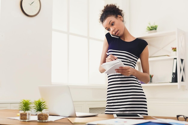 Poważna afro-amerykańska kobieta biznesu w pracy rozmawia przez telefon i robi notatki, stojąc w swoim miejscu pracy w biurze, kopia przestrzeń