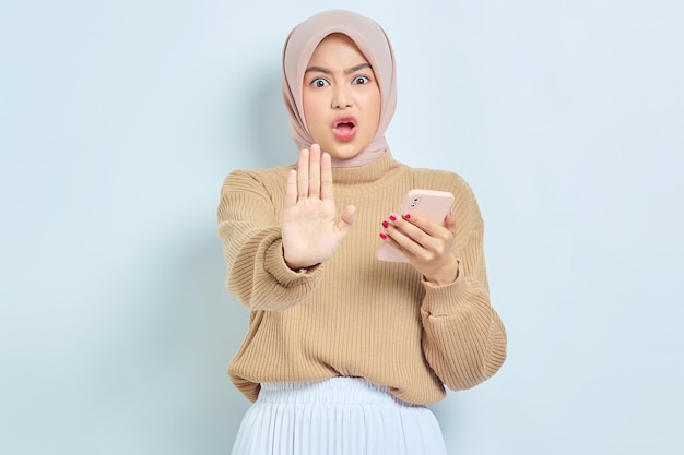 Poważna azjatycka muzułmańska kobieta w brązowym swetrze stojąca z wyciągniętą ręką pokazując gest zatrzymania na białym tle na białym tle