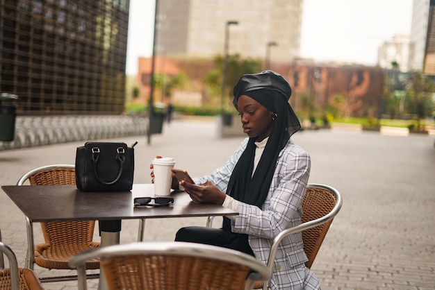 Poważna czarna kobieta za pomocą smartfona przy stole w kawiarni na świeżym powietrzu